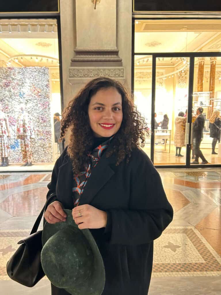 Femme souriante dans une galerie ornée lors d'un week-end à Milan avec des boutiques en arrière-plan.