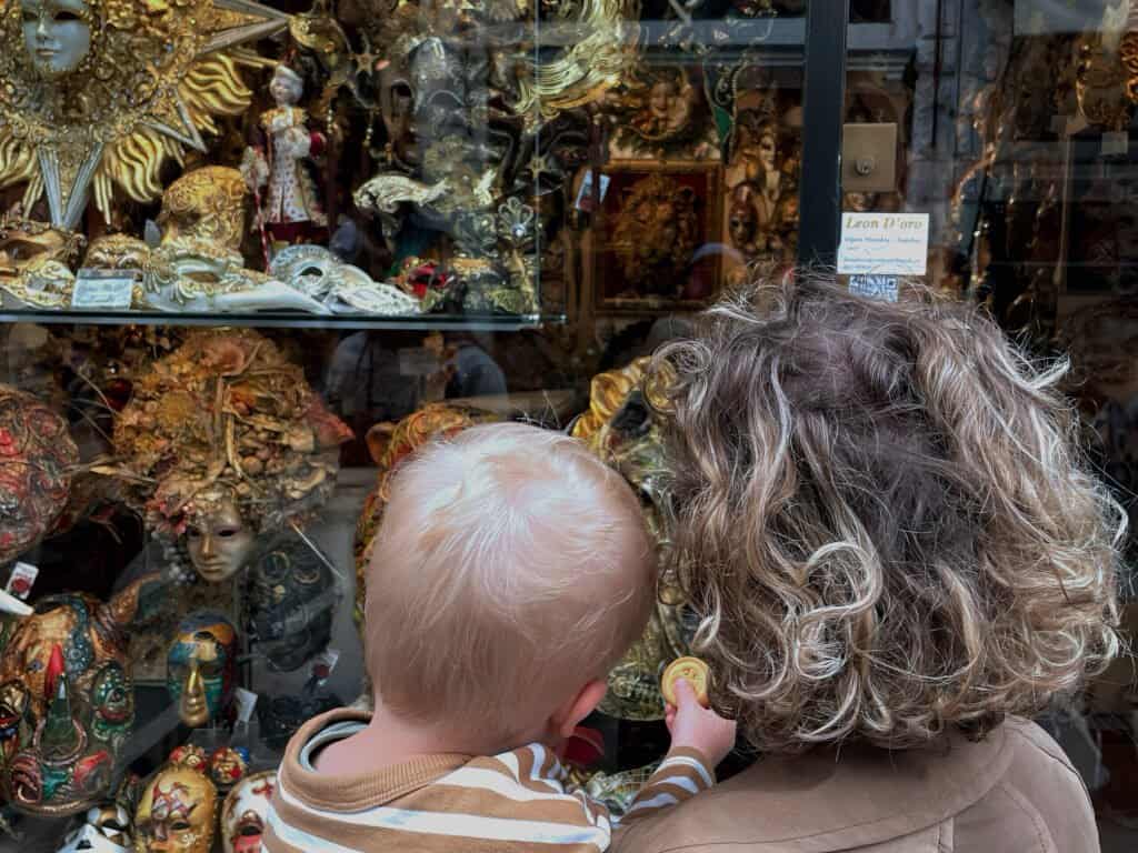 Un enfant et un adulte aux cheveux bouclés regardent une vitrine ornée de masques vénitiens colorés et d'objets décoratifs détaillés. En explorant Venise avec des enfants, l'enfant montre avec enthousiasme l'un des masques, enchanté par ses couleurs vibrantes et ses motifs complexes.