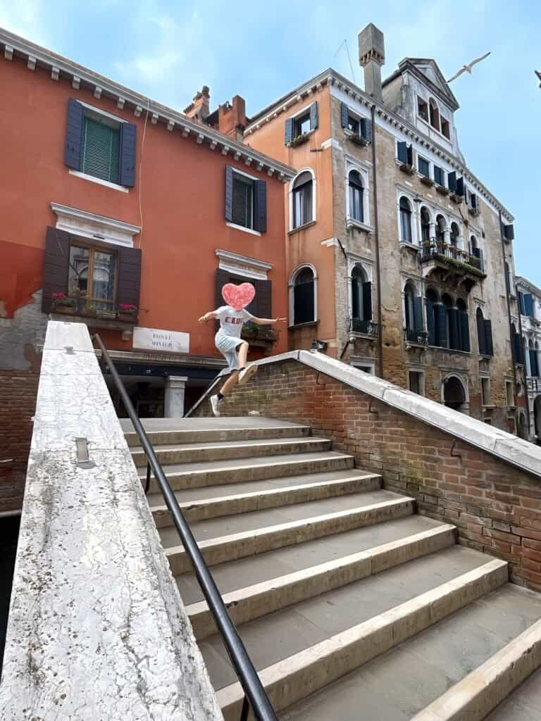 Personne en tenue décontractée tenant une pancarte en forme de cœur tout en sautant sur un pont de pierre à Venise avec des enfants, sur fond de bâtiments historiques colorés.