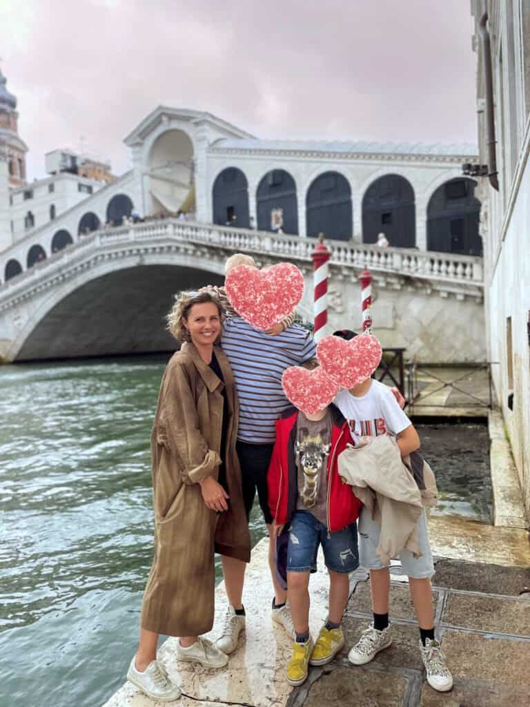 Une famille de quatre personnes se tient au bord d'un canal à Venise avec des enfants, en arrière-plan le pont du Rialto. Les adultes sourient tandis que les visages des enfants sont cachés par des autocollants en forme de cœur.