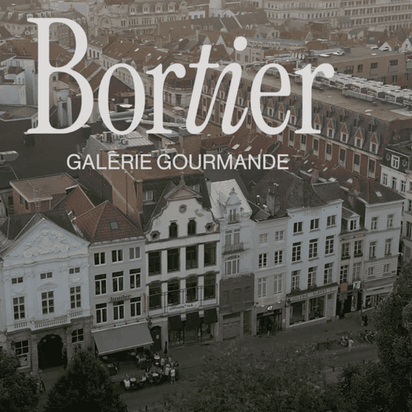 Vue aérienne d'une rue européenne de Bruxelles avec des bâtiments historiques. Le texte superposé indique « Bortier Galerie Gourmande », faisant allusion au marché alimentaire animé et à la gastronomie riche qui définissent le charme de la ville.