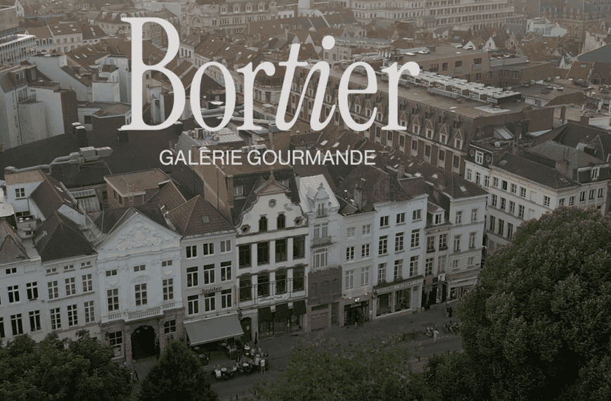 Vue aérienne d'une rue européenne de Bruxelles avec des bâtiments historiques. Le texte superposé indique « Bortier Galerie Gourmande », faisant allusion au marché alimentaire animé et à la gastronomie riche qui définissent le charme de la ville.